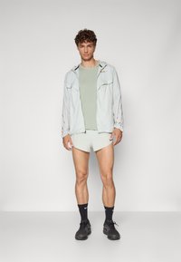 Chaqueta ligera de color gris pálido con detalles en negro y dorado, camiseta verde y pantalones cortos claros. El modelo lleva zapatillas negras y calcetines.