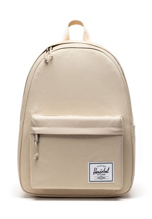 Sac à dos beige Herschel avec poche zippée à l'avant et écusson logo rectangulaire blanc, doté d'une poignée supérieure et de deux tirettes de fermeture éclair.