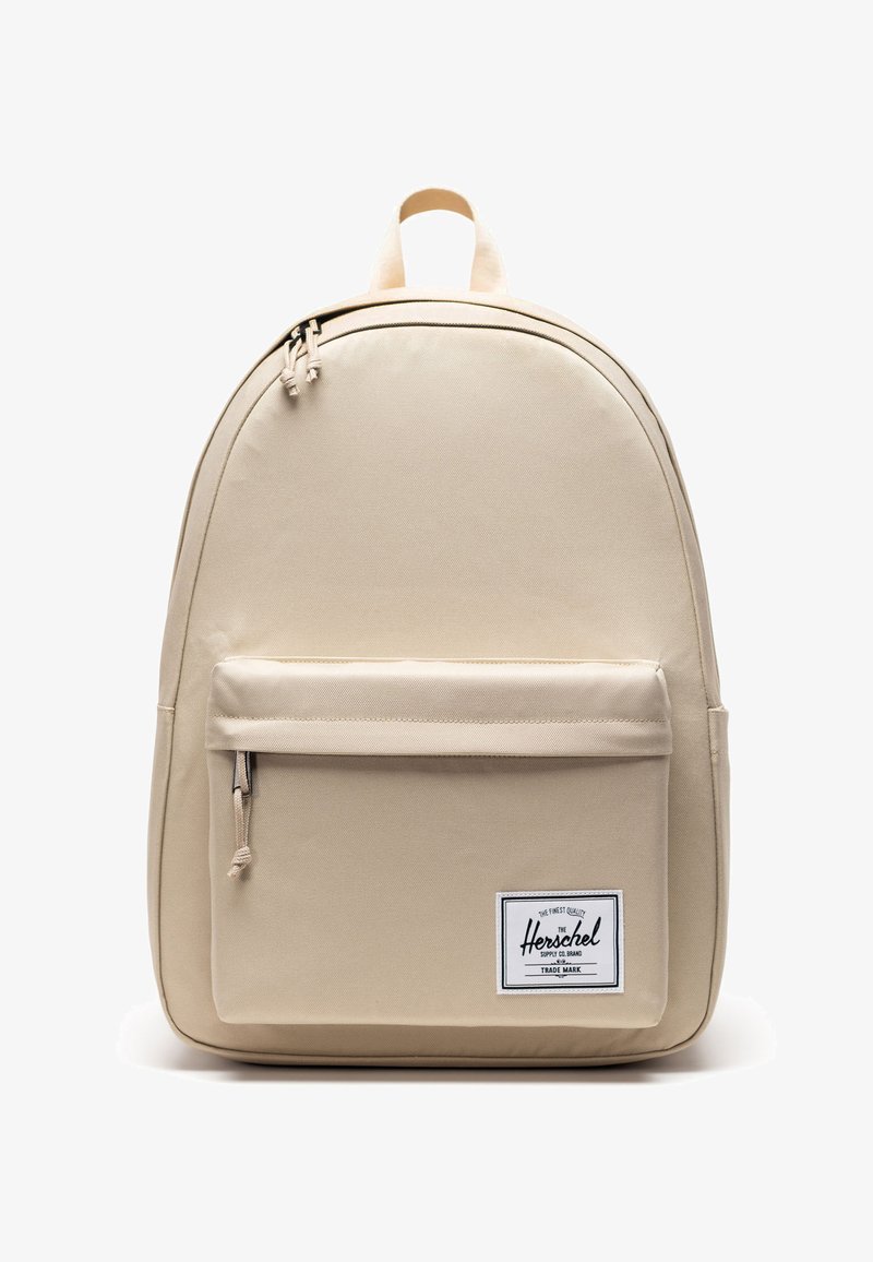 Sac à dos beige Herschel avec poche zippée à l'avant et écusson logo rectangulaire blanc, doté d'une poignée supérieure et de deux tirettes de fermeture éclair.