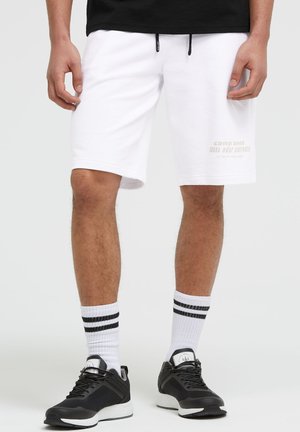 Mann trägt weiße knielange Shorts, schwarze Sneaker, weiße Socken mit zwei schwarzen Streifen und ein schwarzes Shirt, steht vor hellem Hintergrund.