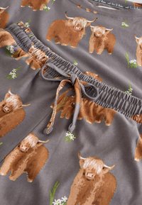 Gråt stof med et gentaget mønster af brune højlandskvæg og hvide blomster. Taljebåndet har elastik og en snøre.