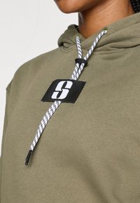 Olijfgroene hoodie van textuurstof, met een zwart logo-patch, witte koord met zwarte accenten en een strakke pasvorm.