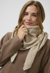 Beige uldhalstørklæde bundet løst om halsen, struktureret med en blød finish. Båret med en brun frakke, der viser en elegant drapering.