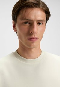 T-shirt leggero di colore beige chiaro con collo a giroclassico, texture liscia e dettagli di cucitura minimali, indossato da un individuo.