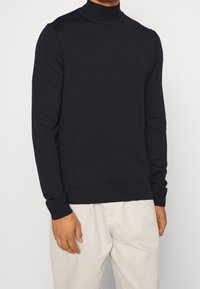 Suéter de cuello alto azul marino con mangas largas, cuellos y puños acanalados, hecho de una tela suave, combinado con pantalones de color claro.