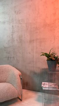 Βελούδινη μπεζ πολυθρόνα δίπλα σε μεταλλικό τραπεζάκι με συρμάτινο πλαίσιο που κρατάει περιοδικά και ένα γλαστράκι, τοποθετημένη μπροστά από υφή τσιμεντένινος τοίχου.