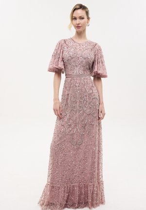 Mujer que lleva un vestido largo rosa con un elaborado bordado de cuentas plateadas y mangas cortas acampanadas, posando sobre un fondo blanco liso.