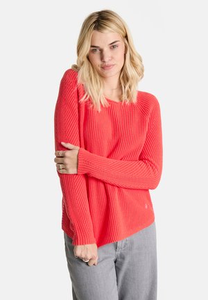 Heller korallenfarbener Strickpullover mit gerippter Textur, langen Ärmeln und lockerer Passform. Kombiniert mit hellgrauen Hosen.