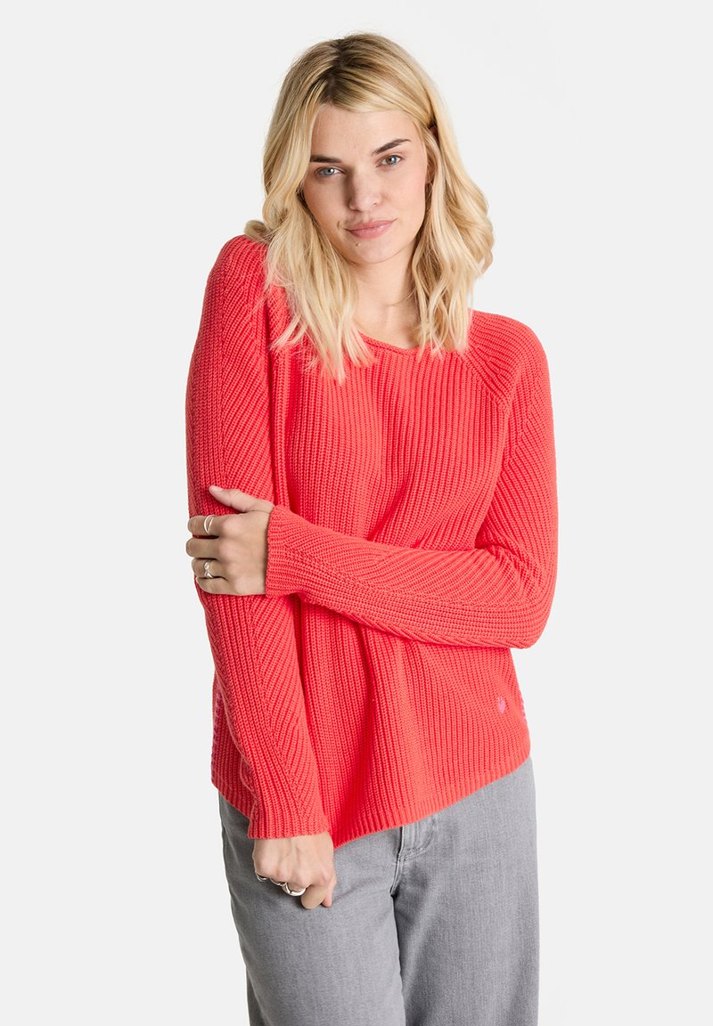 Heller korallenfarbener Strickpullover mit gerippter Textur, langen Ärmeln und lockerer Passform. Kombiniert mit hellgrauen Hosen.