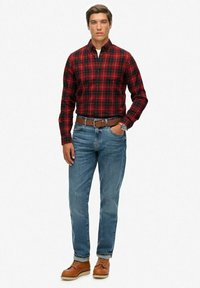 Rood en zwart geruite shirt, blauwe spijkerbroek, bruine riem en tan schoenen. Model poseert met handen in de zakken, tegen een effen witte achtergrond.