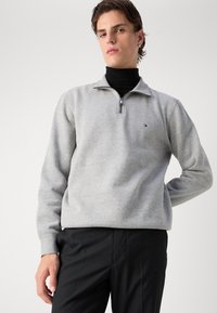 Šviesiai pilkas zip-up džemperis iš minkštos medžiagos, su aukštu apykakle ir siuvinėtu logotipu kairiajame krūtinės kampe. Juodos kelnės užbaigia įvaizdį.