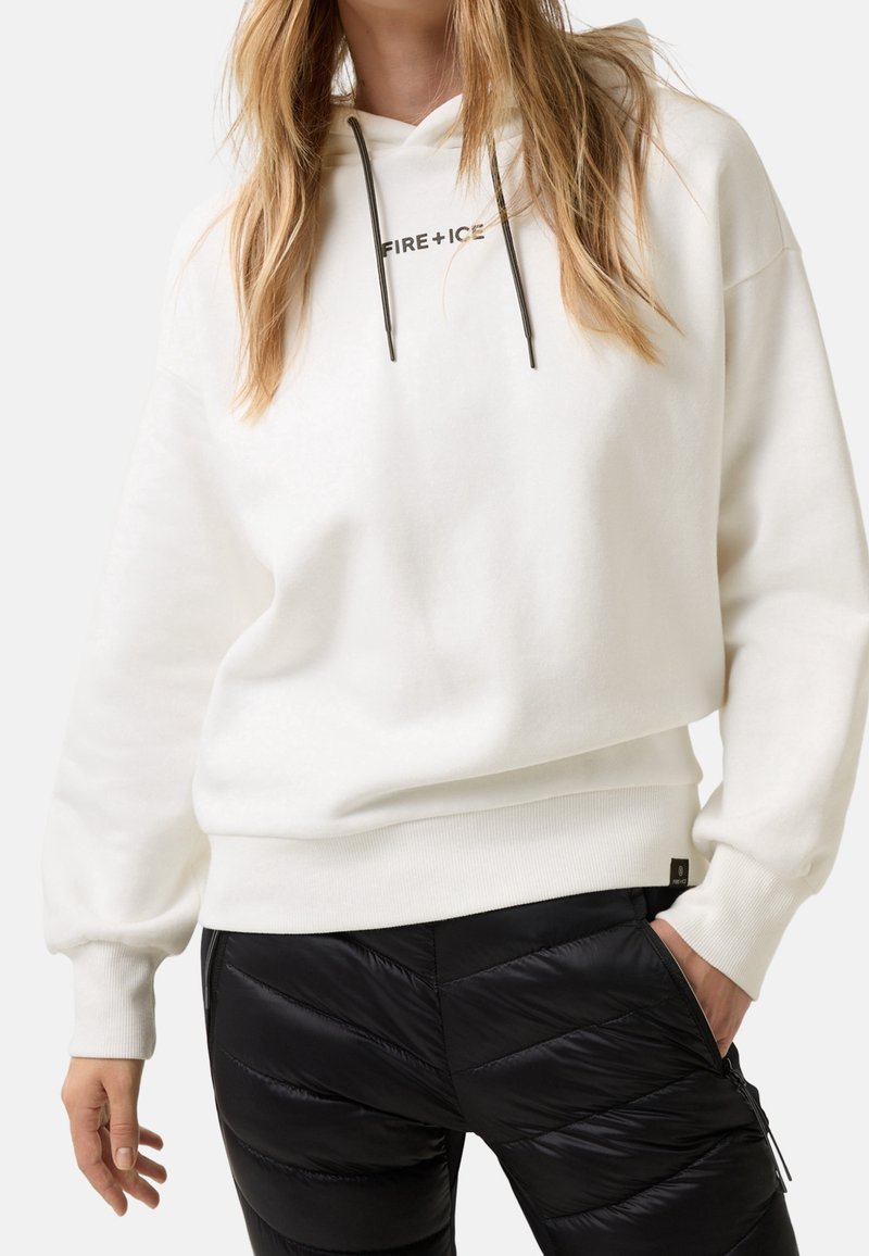 Femme portant un sweat à capuche blanc avec le texte "FIRE + ICE" et un pantalon matelassé noir, une main dans la poche, sur un fond clair uni.