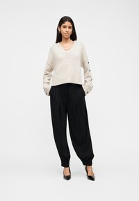 Femme portant un pull beige à col en V avec des boutons aux manches, un pantalon noir plissé et fuselé, des talons noirs, et un collier de perles, debout les mains dans les poches.