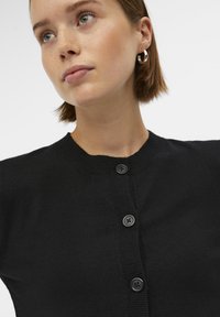 Čierny pletený kardigán s okrúhlym výstrihom, so zapínaním na gombíky vpredu a rebrovanými manžetami, predvedený na modeli na bielom pozadí.
