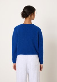 Sieviete ar tumšiem cirtainiem matiem bulciņā ģērbjusies zilā pūkainā džemperī un gaiši pelēkās biksēs, stāvot pret vienkāršu gaišu fonu.