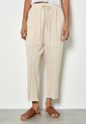 Beżowe lniane spodnie z pionowymi oczkami, elastycznym pasem, troczkiem, podwiniętymi nogawkami, noszone z brązowymi klapkami i białym szydełkowanym topem.
