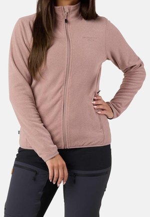 Femme portant une veste polaire zippée mauve clair avec un pantalon d'extérieur noir, debout avec une main sur la hanche et l’autre détendue.