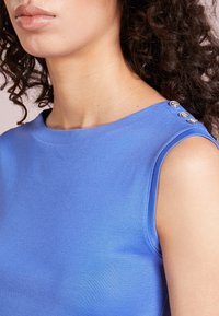 Blå ärmlös topp gjord av ett mjukt tyg. Har en rund halsringning och tre dekorativa knappar på axeln.