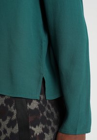 Detailní záběr na zelenou blůzu s dlouhými rukávy a bočním rozparkem, která je kombinována se vzorovanou sukní s tmavými a kovovými odstíny.