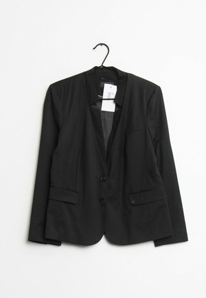 Blazer noir sur mesure avec col à revers, deux boutons, poches à rabat à l'avant, suspendu sur un cintre noir avec une étiquette de prix blanche.