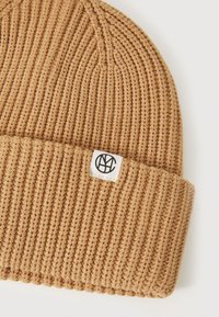 Knit beanie in tan color with a ribbed texture. Features a folded cuff with a small white label displaying a logo.