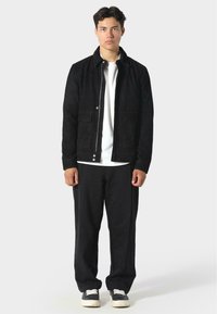 Černá semišová bunda se zapínáním na zip vpředu, se dvěma dolními kapsami a klasickým límcem, nošena přes bílou košili a černé kalhoty, doplněná černými teniskami.