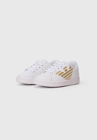 Hvit lave sneakers med gullørnlogo på sidene og hvite lisser, vinklet for å vise detaljer på fronten og ytre siden.