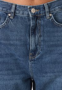 Modré džínové kalhoty s vysokým pasem, stříbrnými kovovými knoflíky a kontrastním žlutým prošíváním na kapsách a švech. Bez viditelného opotřebení.