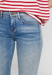 Vaqueros azules deslavados con un corte ajustado, que cuentan con un diseño clásico de cinco bolsillos y cierre de botón. Combinados con una blusa de rayas rojas y blancas.