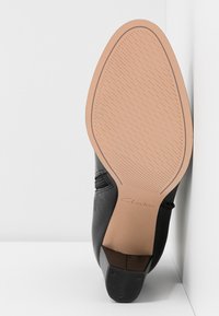 Semelle d'une chaussure en cuir noir avec une semelle extérieure texturée marron clair et un petit talon noir, positionnée à la verticale contre une surface blanche.