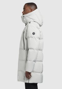 Világosszürke puffKabát magas gallérral, cipzáras zsebekkel és logóval ellátott vállrésszel. Texturált, steppelt kialakítás jellemzi.
