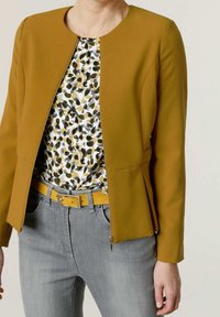 Femme portant une veste cintrée jaune moutarde sur un chemisier blanc à motif floral, combinée à un jean gris et une ceinture jaune assortie.