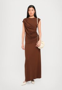 Brun drapert maxi-kjole med kappeärmer, laget av myk stoff. Antrekket inkluderer en kremfarget clutch og lyse spisse sko.