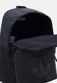 Musta kangaskassi, jossa on ylhäältä suljettava vetoketju, tilava sisätila ja etutasku, jossa on Adidas-logo. Sileä rakenne ja minimalistinen muotoilu.