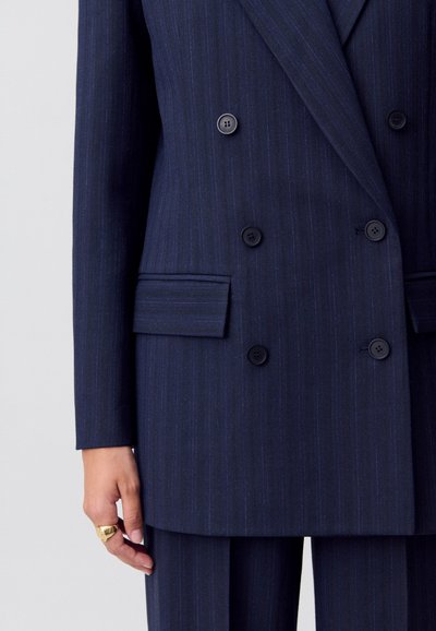 Blazer azul marino de doble botonadura con rayas finas, grandes solapas y botones negros. Presenta un bolsillo de ojal y una textura suave.