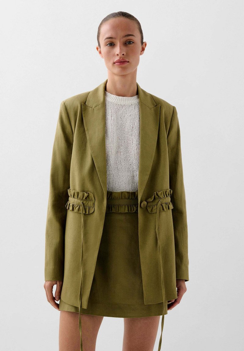 Olive green linen blazer with ruffled side accents, paired with a matching skirt, and a light textured sweater beneath. Simple, tailored design.