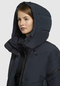 Pufjakke i mørk navy med en stor, struktureret hætte. Har en lynlåslukning, trykknapper og en sidelomme med et elegant accent.