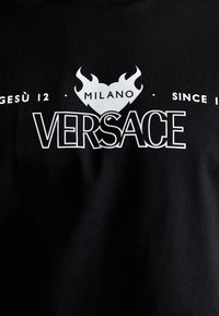 Must musta T-särk, millel on valget värvi trükitud graafika, sealhulgas "VERSACE," leegid ja "MILANO" tekst, luues julge kontrasti.