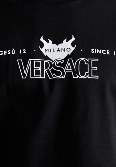 Juodas medvilninis marškinėliai su baltais spaudiniais, įskaitant "VERSACE", liepsnas ir tekstą "MILANO", sukuriantys drąsų kontrastą.