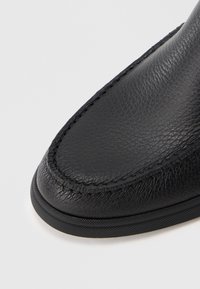 Zapato de cuero negro con una superficie texturizada, punta redondeada y costuras visibles en el borde. Suela de goma duradera con un acabado suave.