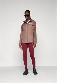 Brown waterproof jacket with a zippered front, logo on chest; paired with maroon leggings and black sneakers. Model wearing sunglasses.