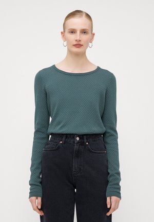 Teksturert langermet topp i petrol med rund hals, parret med høy midje svarte jeans. Utstyrt med ribbede mansjetter og subtilt mønster.