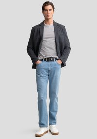Blazer gris sobre una camiseta gris, combinado con jeans azul claro. Zapatillas blancas con suelas marrones. El modelo está de pie con las manos en los bolsillos.
