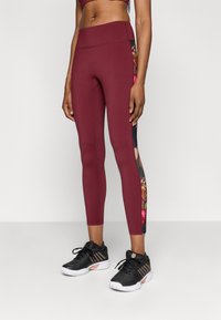 Bordó leggings magas derékkal, oldalon virágmintával; fekete sportcipővel párosítva. Sima szövet textúra.