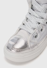 Zapatilla alta de color gris claro con parte superior acolchada, puntera plateada brillante, cordones reflectantes y suela de goma blanca texturizada.