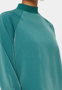 Maglia a manica lunga turchese con colletto alto, texture liscia e maniche raglan. Il tessuto sembra leggero e leggermente lucido.