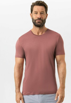 Bärtiger Mann, der ein eng anliegendes kurzärmliges T-Shirt in staubigem Rosaton und vertikal gestreifte Hosen trägt, steht vor einem schlichten weißen Hintergrund.