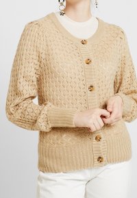 Cardigan en maille beige avec un motif texturé, col rond et manches longues. Comprend cinq boutons marron sur le devant.