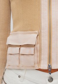 Μπεζ ριμπ πλεκτό πουλόβερ με κεντρικό φερμουάρ, που διαθέτει μια ανοιχτόχρωμη τσέπη με δύο καλύμματα και διακοσμητικές ραφές. Απαλή υφή και εφαρμοστή σχεδίαση.