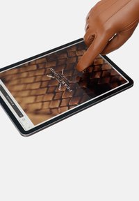 Tablet med sort ramme, der viser et struktureret mønster. En brun læderhandske peger på skærmen og fremhæver et brandlogo.